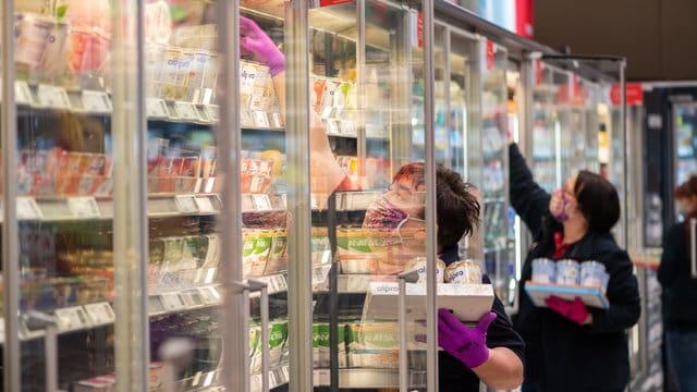 Mitarbeiterinnen in einem Supermarkt tragen Schutzmasken, während sie die Kühlregale mit Waren bestücken. Mitarbeiterinnen in einem Supermarkt tragen Schutzmasken, während sie die Kühlregale mit Waren bestücken.