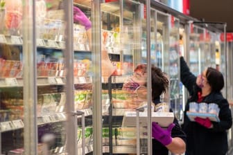 Mitarbeiterinnen in einem Supermarkt tragen Schutzmasken, während sie die Kühlregale mit Waren bestücken.
