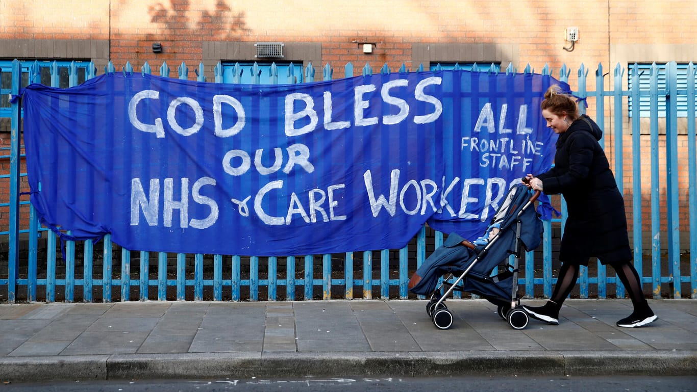 Eine Frau mit Kinderwagen läuft an einem Unterstützungsbanner für die Mitarbeiter des NHS vorbei. Auf dem Banner steht auf Englisch: "Gott segne unsere NHS-Pflegekräfte."