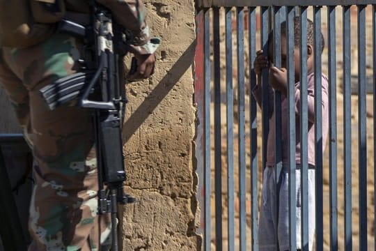 Ein Kind hält sich im südafrikanischen Johannesburg an einem Torgitter fest, hinter dem ein Mitglied der Nationalen Verteidigungskräfte steht.