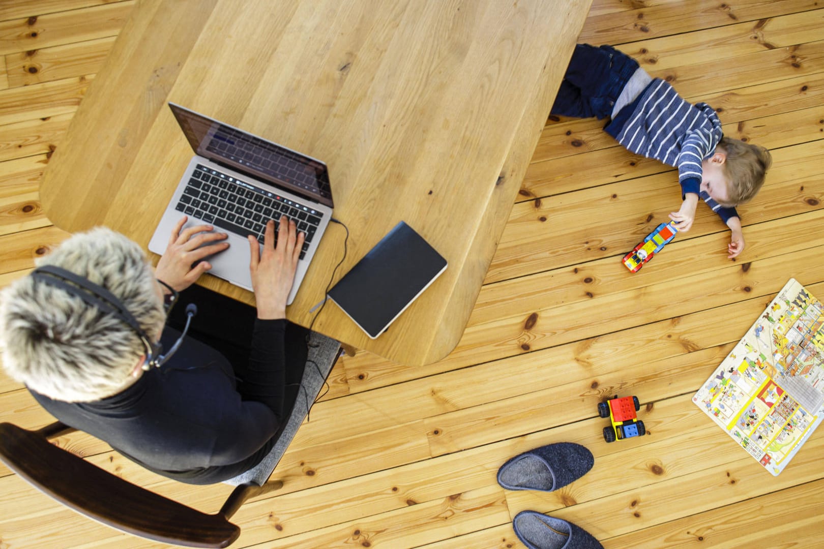 Eine Frau arbeitet im Homeoffice, während ihr Kind unter dem Tisch spielt (Symbolfoto): Viele Eltern müssen ihre Kinder derzeit zu Hause betreuen.