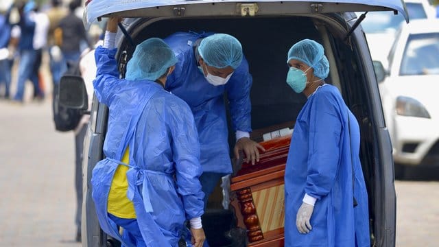 Männer mit Schutzanzügen laden vor einem Krankenhaus in Ecuador einen Sarg in einen Wagen. Männer mit Schutzanzügen laden vor einem Krankenhaus in Ecuador einen Sarg in einen Wagen.