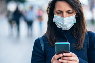 Eine Frau mit Maske und Smartphone: Bei der Corona-Warn-App sollen die Daten dezentral gespeichert werden.