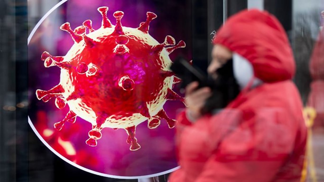 Eine elektronische Werbetafel zeigt eine Illustration des Coronavirus, davor steht ein Mensch mit seinem Smartphone: Das Virus werde uns auch 2021 beschäftigen, so ein Münchner Arzt.