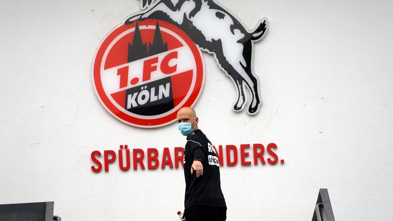 Physiotherapeut Christian Osebold vom 1. FC Köln steht mit Mundschutz vor einem Klub-Logo: In der Corona-Krise unterstützt der Bundesligist das Erzbistum Köln.