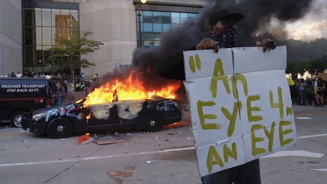 Ein Demonstrant hält bei einer Protestaktion in der Nähe eines brennenden Polizeiautos ein Schild mit dem Anfang eines Zitats aus der Bibel "Auge um Auge". Ein Demonstrant hält bei einer Protestaktion in der Nähe eines brennenden Polizeiautos ein Schild mit dem Anfang eines Zitats aus der Bibel "Auge um Auge".