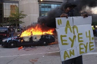 Ein Demonstrant hält bei einer Protestaktion in der Nähe eines brennenden Polizeiautos ein Schild mit dem Anfang eines Zitats aus der Bibel "Auge um Auge".
