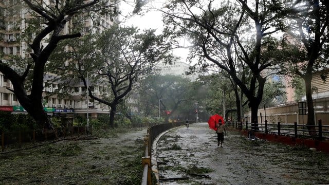 Ein Mann mit einem Regenschirm geht über eine Straße in Mumbai, auf der vom Wind abgerissene Äste und Blätter liegen. Ein Mann mit einem Regenschirm geht über eine Straße in Mumbai, auf der vom Wind abgerissene Äste und Blätter liegen.