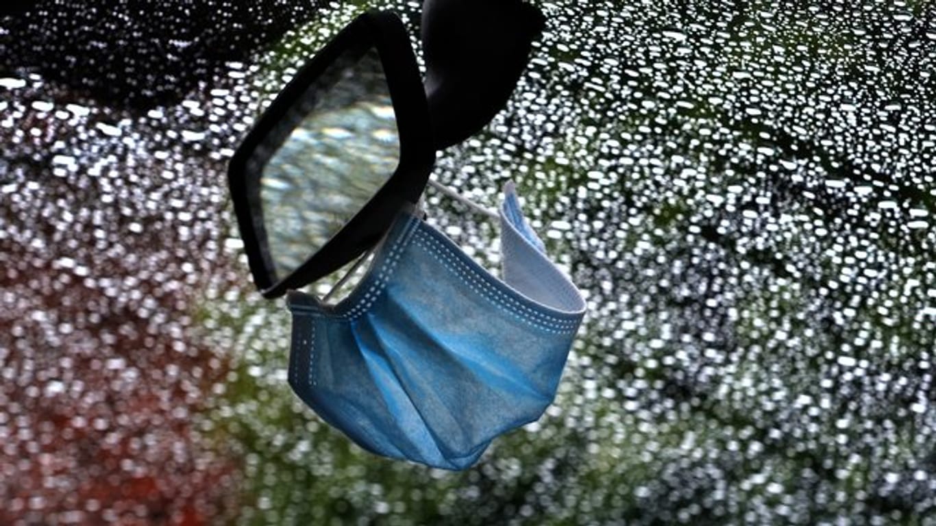 Ein medizinischer Mundschutz am Rückspiegel eines im Regen parkenden Autos.