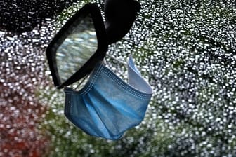 Ein medizinischer Mundschutz am Rückspiegel eines im Regen parkenden Autos.
