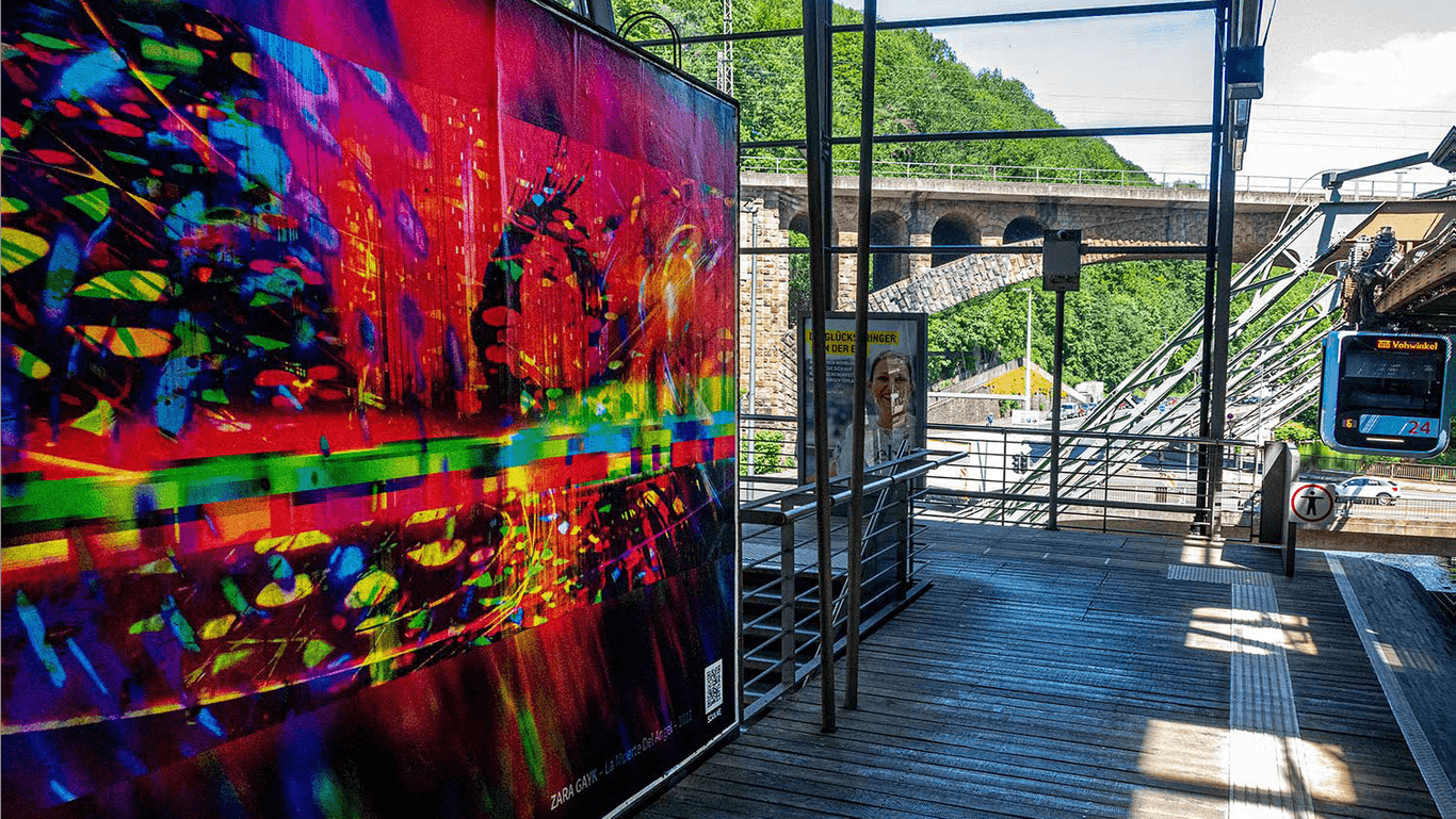 Ein Plakat des Künstlers Zara Gayk hängt an der Schwebebahnstation am Zoo/Stadion in Wuppertal: Über 150 solcher Motive sind in der ganzen Stadt zu sehen.