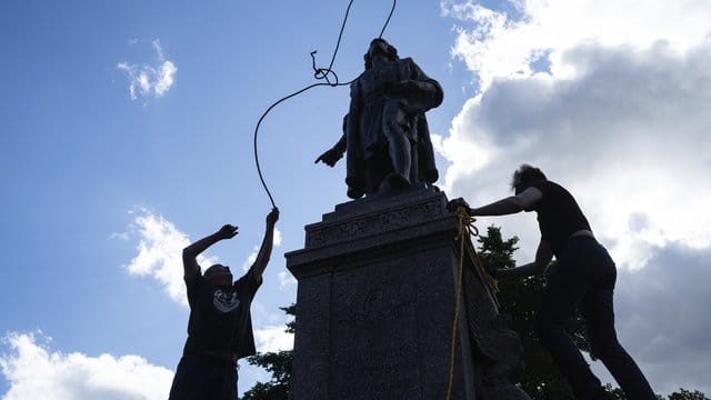 Zwei Männer befestigen ein Seil um den Hals einer Statue von Christopher Columbus. Zwei Männer befestigen ein Seil um den Hals einer Statue von Christopher Columbus.