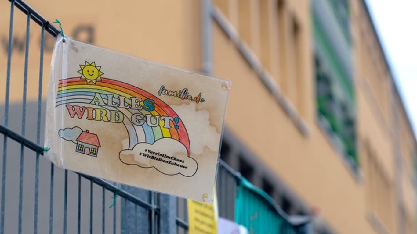 Ein Schild mit den Aufschriften "Alles wird gut!" und "#VereintImChaos" sowie "#WirBleibenZuhause" hängt am Zaun einer Grundschule.