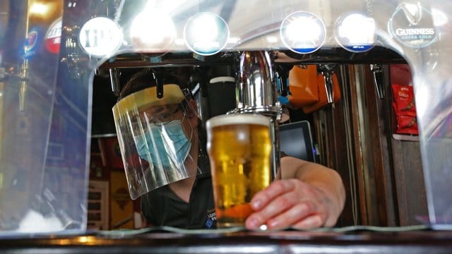 Ein Barkeeper im Pub zapft ein Glas Bier mit Mundschutz und Visier vor der Wiedereröffnung. Ein Barkeeper im Pub zapft ein Glas Bier mit Mundschutz und Visier vor der Wiedereröffnung.