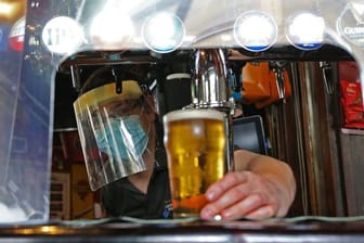 Ein Barkeeper im Pub zapft ein Glas Bier mit Mundschutz und Visier vor der Wiedereröffnung.