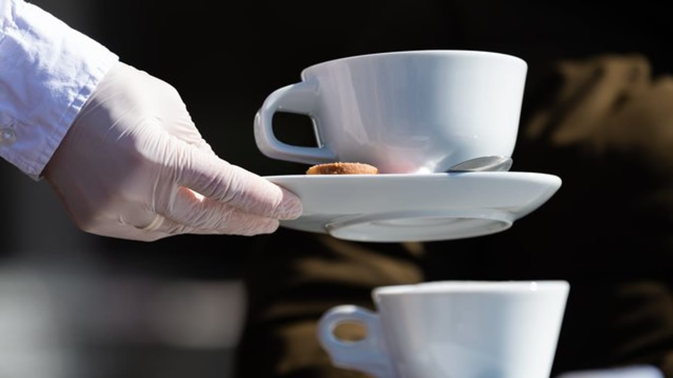Eine Servierkraft bringt Gästen eines Cafés mit Gummihandschuhen Kaffee (Symbolbild): In der Gastronomie ist der Thekenbetrieb für Speisen und Getränke wieder erlaubt.