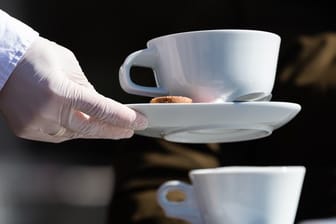 Eine Servierkraft bringt Gästen eines Cafés mit Gummihandschuhen Kaffee (Symbolbild): In der Gastronomie ist der Thekenbetrieb für Speisen und Getränke wieder erlaubt.