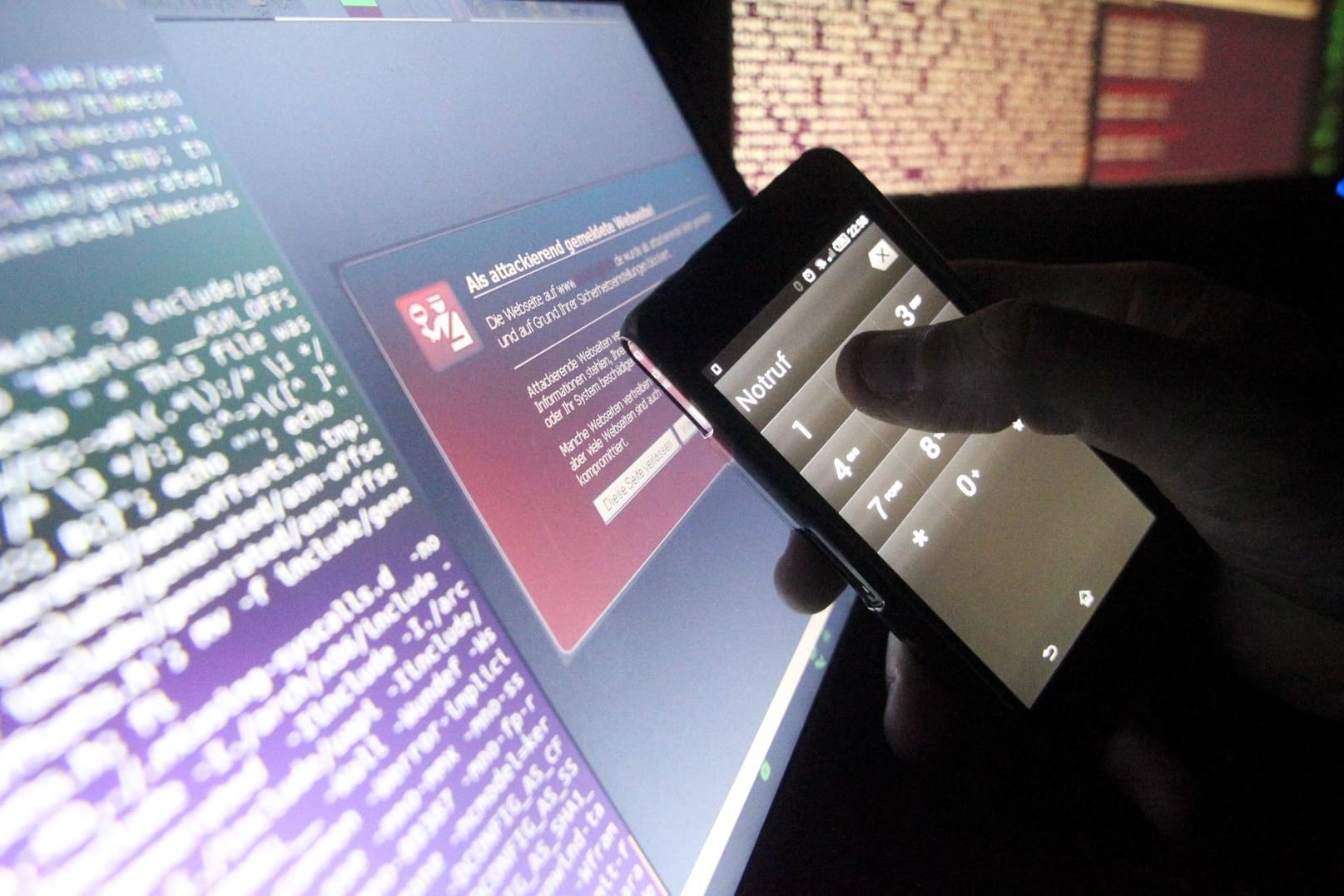 Symbolfoto zum Thema Internetkriminalität: Ein Mann hält ein Smartphone mit Notrufeinstellung vor einem Computermonitor.