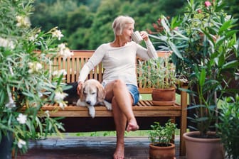 Eine ältere Frau sitzt mit ihrem Hund und einer Tasse Kaffee auf einer Bank im Garten: Um einen entspannten Ruhestand zu haben, sollten Sie sich Ihre Rentenlücke ausrechnen – und entsprechend Geld sparen.