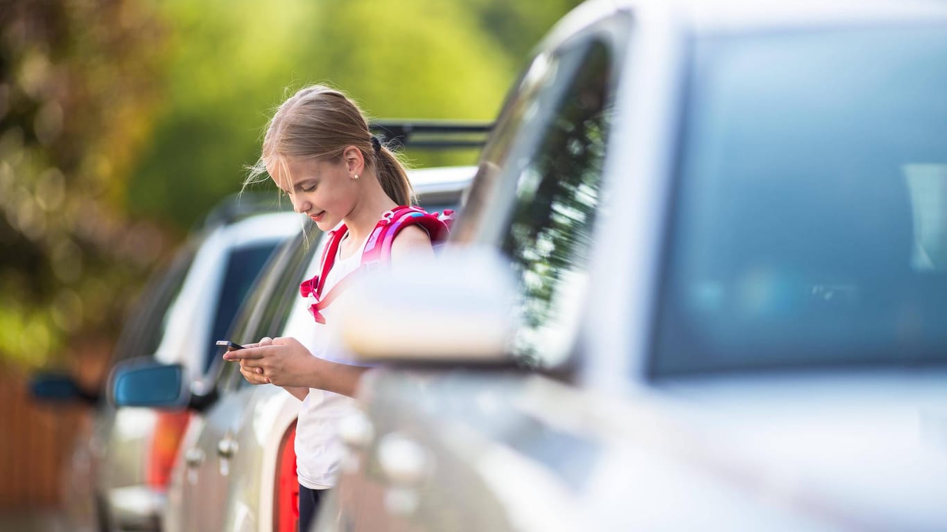 Ein Mädchen steht zwischen parkenden Autos und schaut auf ein Handy (Symbolbild): Zum Schutz Tausender Erstklässler will die Stadt Düsseldorf verstärkt Geschwindigkeitskontrollen durchführen.