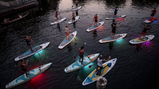 Teilnehmer einer Ausflugsgruppe fahren mit leuchtenden LED-Stand-Up-Paddles in den Abendstunden über einen Alsterfleet in Hamburg.