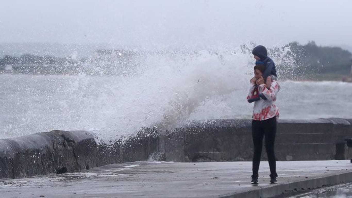 Eine Welle tritt über das Ufer während eine Frau mit einem Kind auf den Schultern an der Strandpromenade geht.