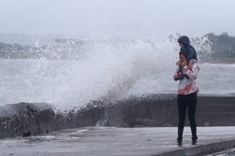 Eine Welle tritt über das Ufer während eine Frau mit einem Kind auf den Schultern an der Strandpromenade geht.