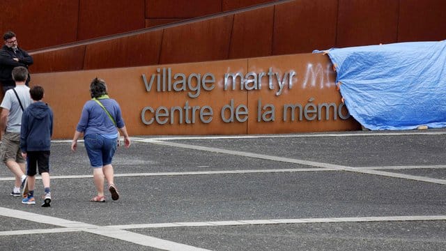 Besucher gehen an einem abgedeckten Graffiti auf dem Schriftzug am Eingang der Gedenkstätte in Oradour-sur-Glane vorbei.