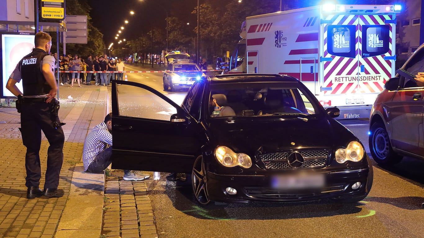 Ein Polizist steht auf einer abgesperrten Straße neben einem schwarzen Mercedes, der in einen Verkehrsunfall verwickelt ist. In und an dem Fahrzeug sitzen zwei Männer, die in dem Auto mitgefahren waren.