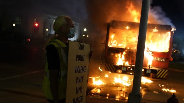 Ein Demonstrant steht mit einem Transparentvor einem brennenden Müllwagen. Ein Demonstrant steht mit einem Transparentvor einem brennenden Müllwagen.