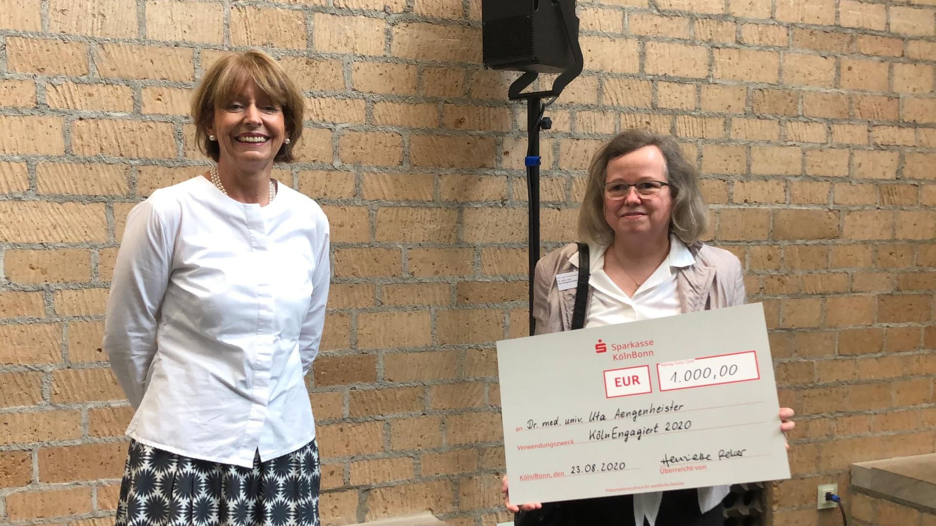 Köln Oberbürgermeisterin Henriette Reker (parteilos) mit Dr. Uta Aengenheister, die ein Schild mit einem Preisgeld in die Höhe hält.