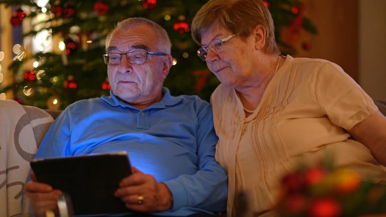 Ein älteres Ehepaar guckt zusammen vor einem geschmückten Tannenbaum in ein Tablet. (Symbolbild)