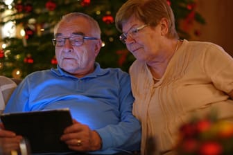 Ein älteres Ehepaar guckt zusammen vor einem geschmückten Tannenbaum in ein Tablet. (Symbolbild)
