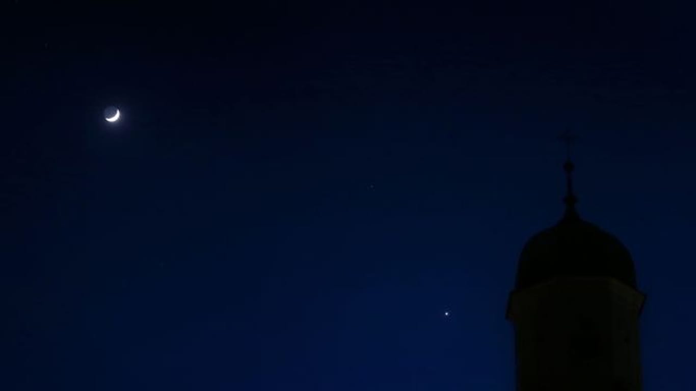 Der zunehmende Mond (l) und der hell leuchtende Planet Venus (r) stehen hinter einem Kirchturm am Abendhimmel.