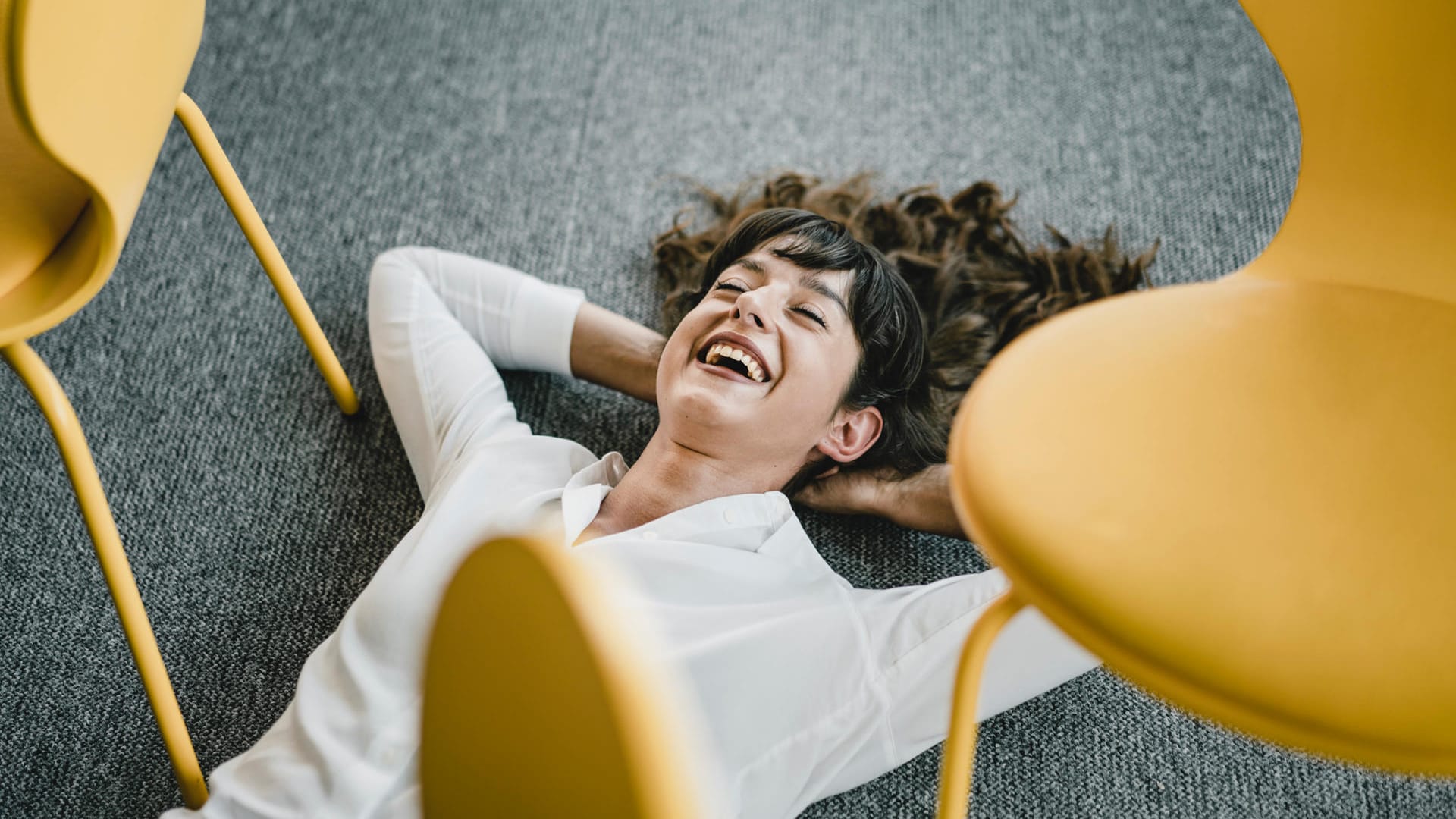 Eine Frau liegt lachend auf dem Boden im Büro: Ob Stress als positiv oder negativ empfunden wird, ist individuell sehr unterschiedlich.