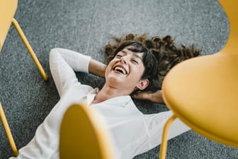 Eine Frau liegt lachend auf dem Boden im Büro: Ob Stress als positiv oder negativ empfunden wird, ist individuell sehr unterschiedlich.