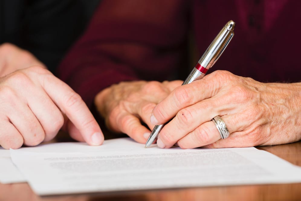 Ältere Frau unterzeichnet ein Dokument: Das Erbrecht garantiert auch ungeliebten Kindern und Verwandten einen Pflichtteil am Nachlass.