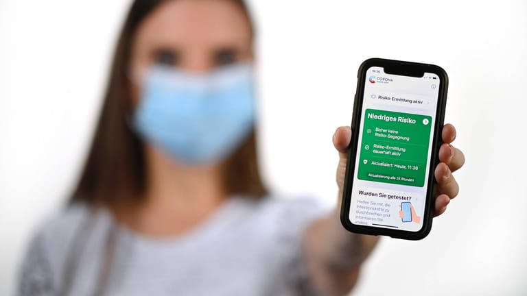 Frau mit Mundschutzmaske, zeigt Smartphone mit Corona Warn-App: Nachverfolgung soll nun auch ohne App funktionieren