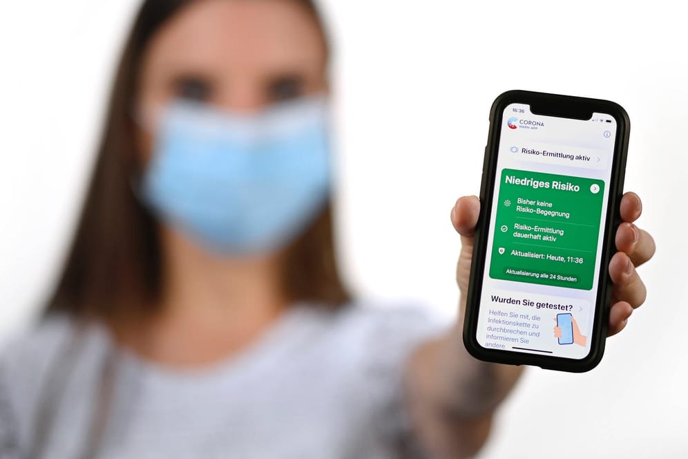 Frau mit Mundschutzmaske, zeigt Smartphone mit Corona Warn-App: Nachverfolgung soll nun auch ohne App funktionieren