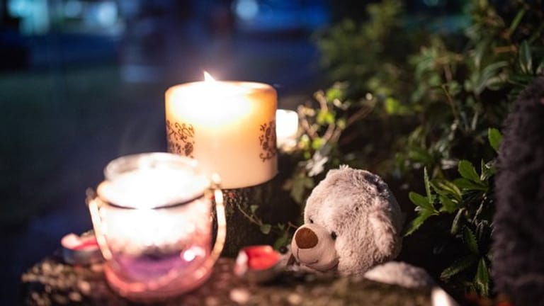 Ein Teddybär und Kerzen liegen und stehen vor einem Haus.