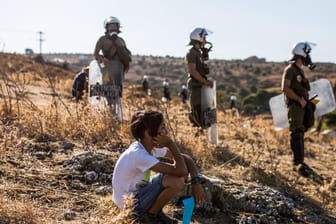 Ein Junge sitzt vor Polizisten und beobachtet einen Protest von Migranten: Auf Lesbos soll ein neues provisorisches Flüchtlingslager aufgebaut werden.