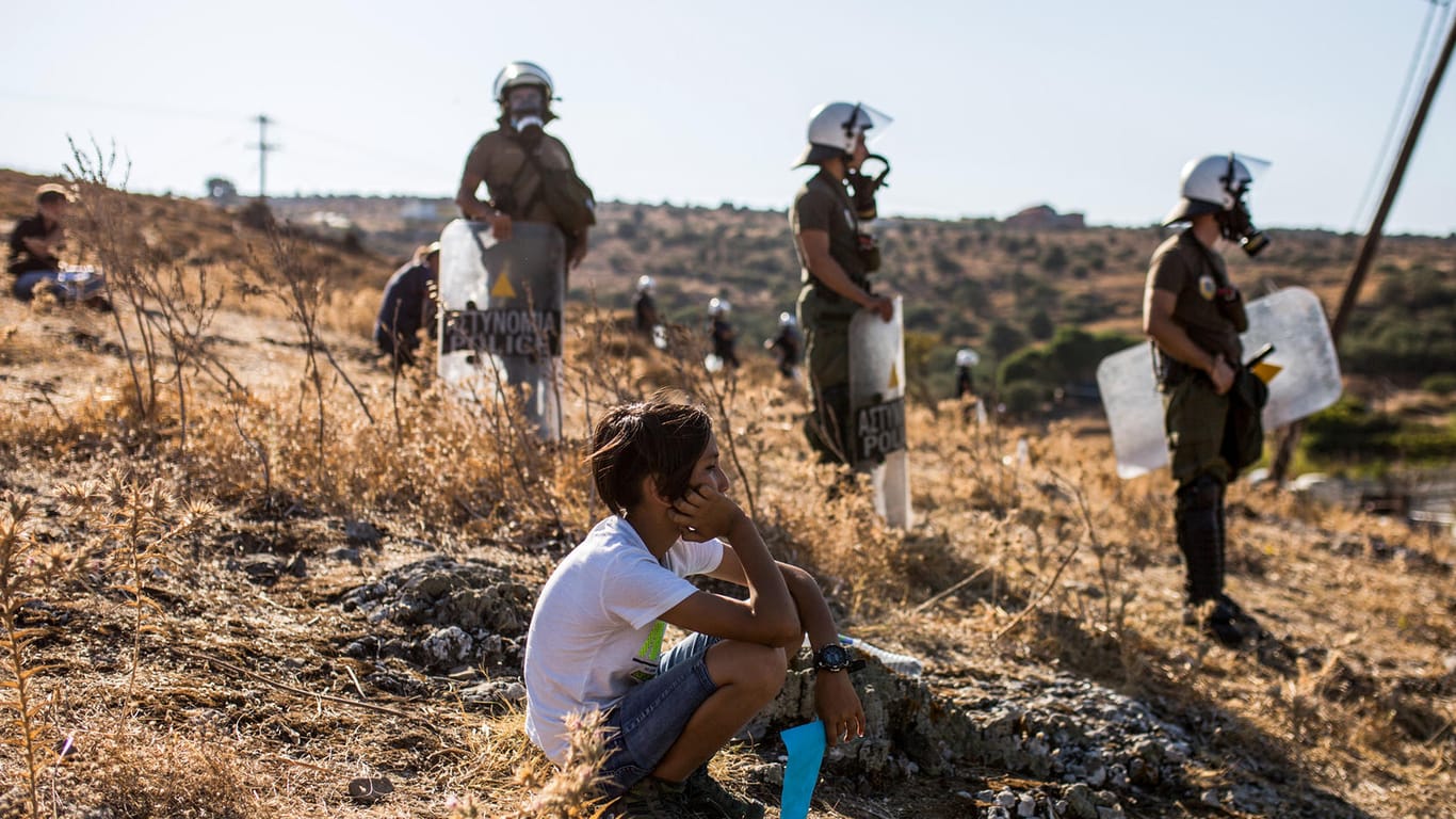 Ein Junge sitzt vor Polizisten und beobachtet einen Protest von Migranten: Auf Lesbos soll ein neues provisorisches Flüchtlingslager aufgebaut werden.