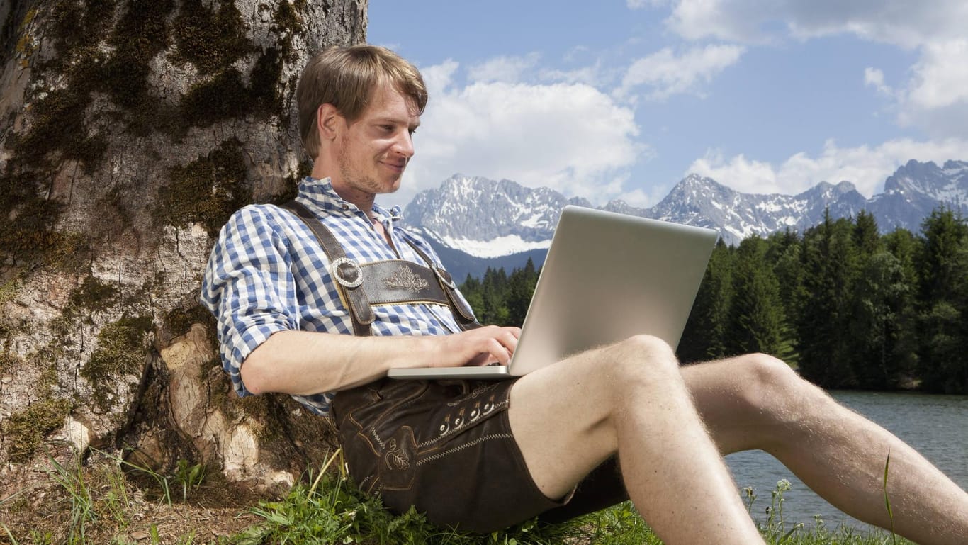 Ein Mann sitzt in Lederhose mit Laptop unter einem Baum (Symbolbild): Eine Initiative ruft Münchner dazu auf, zu Hause Tracht zu tragen.