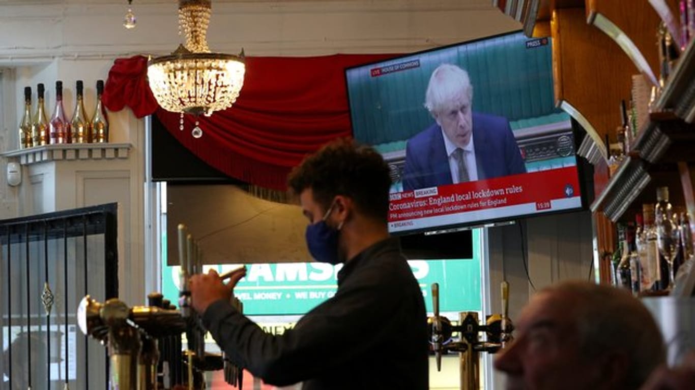 Ein Mitarbeiter eines Liverpooler Pubs schenkt Bier ein, während Premierminister Johnson im Hintergrund das neue Drei-Stufen-System erklärt.