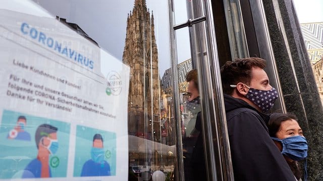 Zwei Personen, die Mund-Nasen-Schutz tragen, stehen in Wien neben einem Hinweisschild für das korrekte Tragen der Mund-Nasen-Bedeckung.