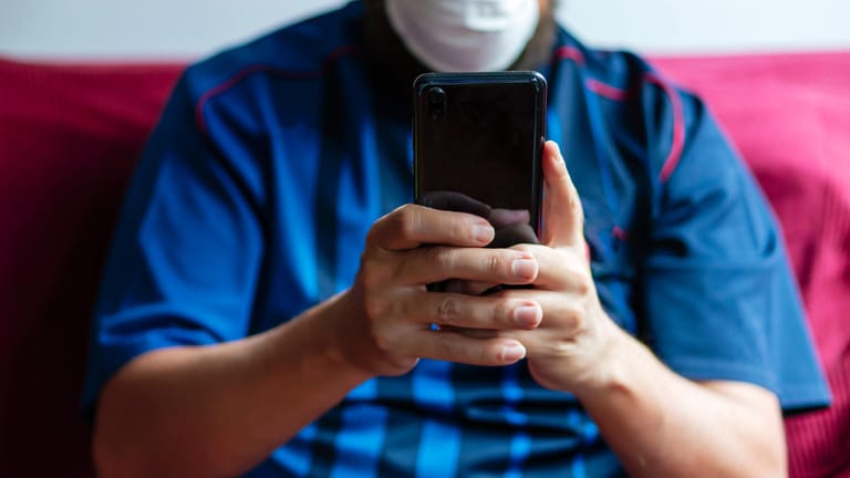 Ein Mann mit Atemschutzmaske nutzt sein Smartphone: Angeblich kann mit Hilfe von Künstlicher Intelligenz ermittelt werden, ob jemand am Coronavirus erkrankt ist.