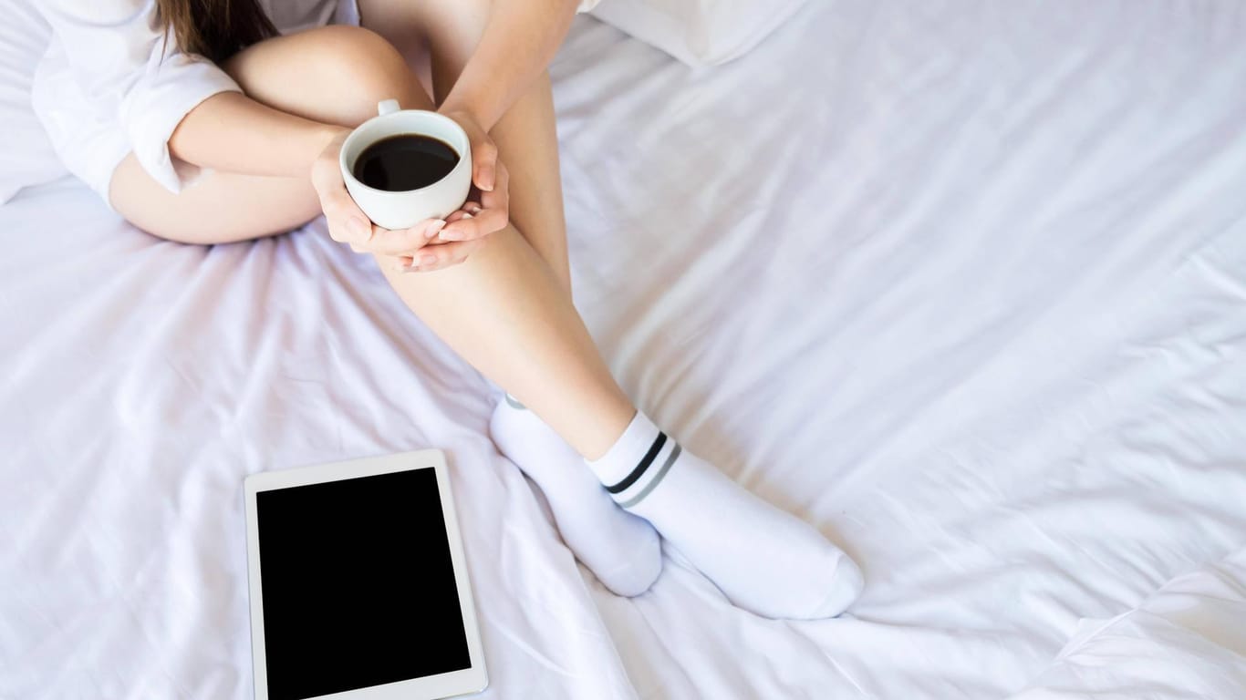 Eine Frau sitzt mit Tablet und Kaffee im Bett (Symbolbild): Im Homeoffice kann man Karneval auch entkommen.