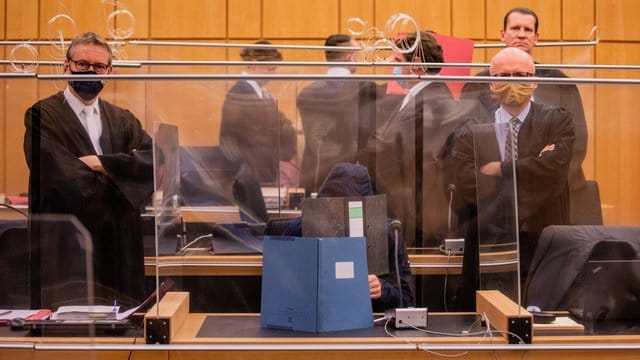 Der 27-Jährige Hauptangeklagte (vorne), seine Mutter (ganz hinten links) und ein dritter Angeklagter (roter Hefter) sitzen in einem Gerichtssaal des Landgerichts.