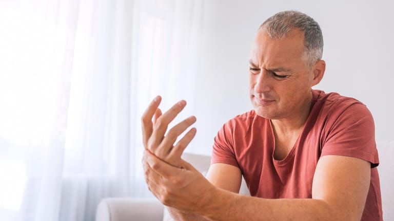 Mann schaut schmerzverkrampft auf seine Hand: Rheuma und weitere Erkrankungen können auch zu einer Osteoporose führen.