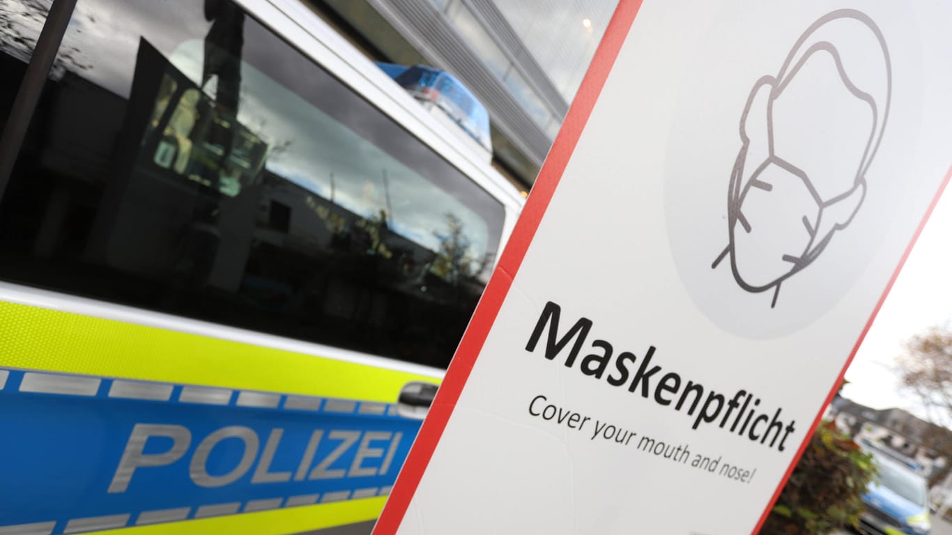 Ein Schild zur Maskenpflicht steht vor einem Polizeiauto (Symbolbild): In Wuppertal haben die Corona-Kontrollen der Stadt bisher Hunderttausende Euro Bußgeld eingebracht.